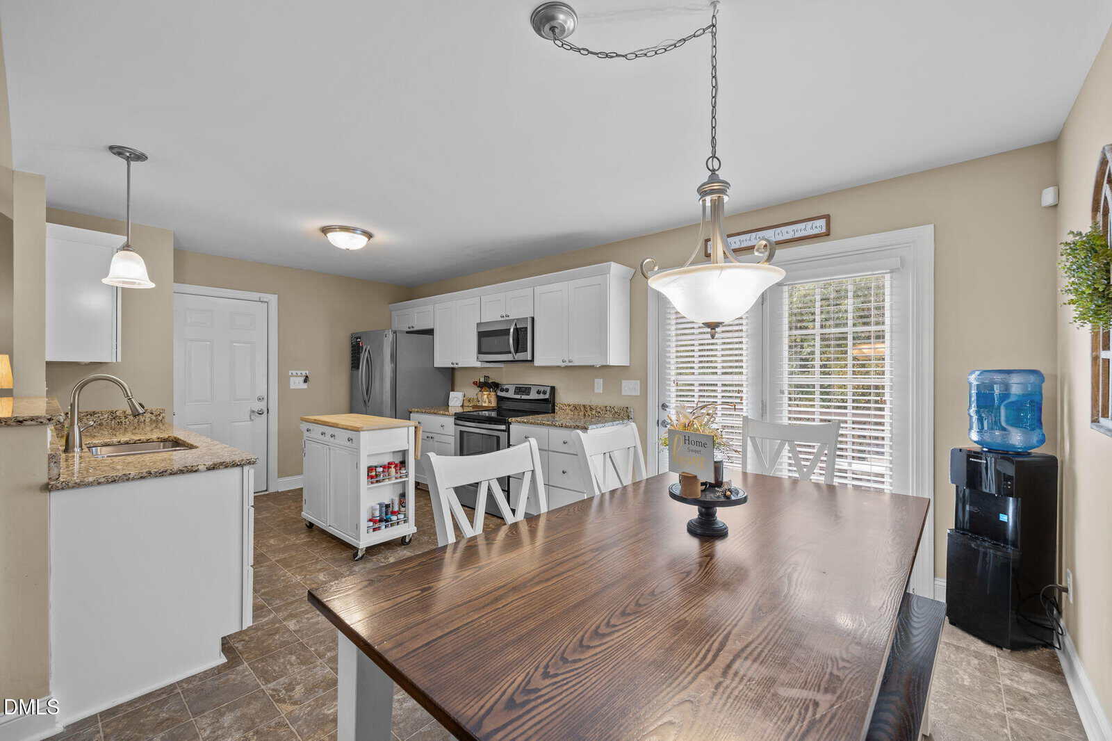 A kitchen with white cabinets, stainless steel appliances, and a wooden dining table with four chairs. There is a water dispenser in the corner and pendant lighting above the table and sink. Sunlight streams in through window blinds.