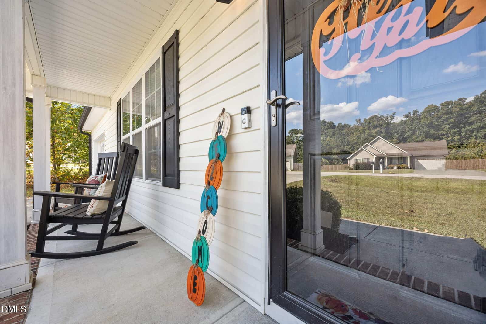 A front porch with a black rocking chair, a stack of decorative pumpkins leaning against the white siding, and a glass door showing the reflection of houses and trees across the street.