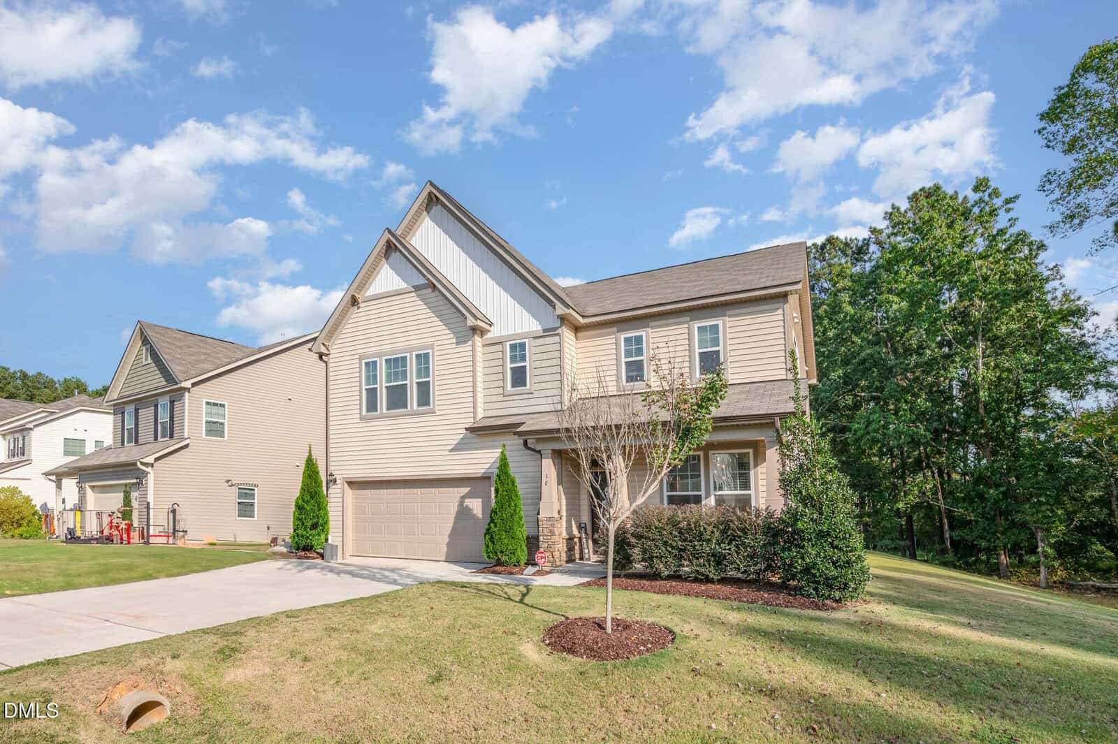 Two-story beige house with a double garage, small front yard, and driveway. The house has white trim, triangular roof peaks, and is surrounded by grass, shrubs, and trees under a partly cloudy sky.
