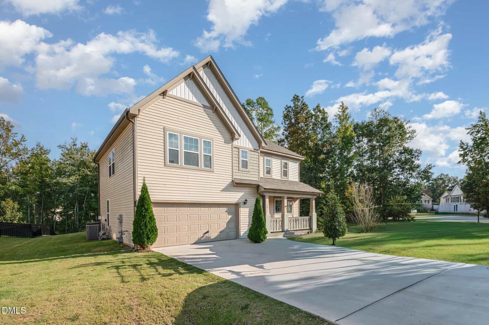 Two-story beige house with a double garage, gabled roof, and small front porch, set on a green lawn with a wide driveway and surrounded by trees, under a blue sky with scattered clouds.