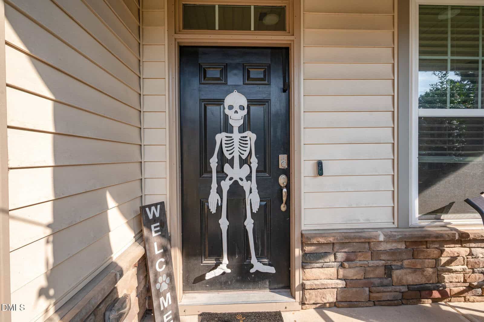 A black front door with a large white skeleton decoration. Next to the door is a black “WELCOME” sign. The house features tan siding and stone accents. Sunlight casts shadows across the porch area.