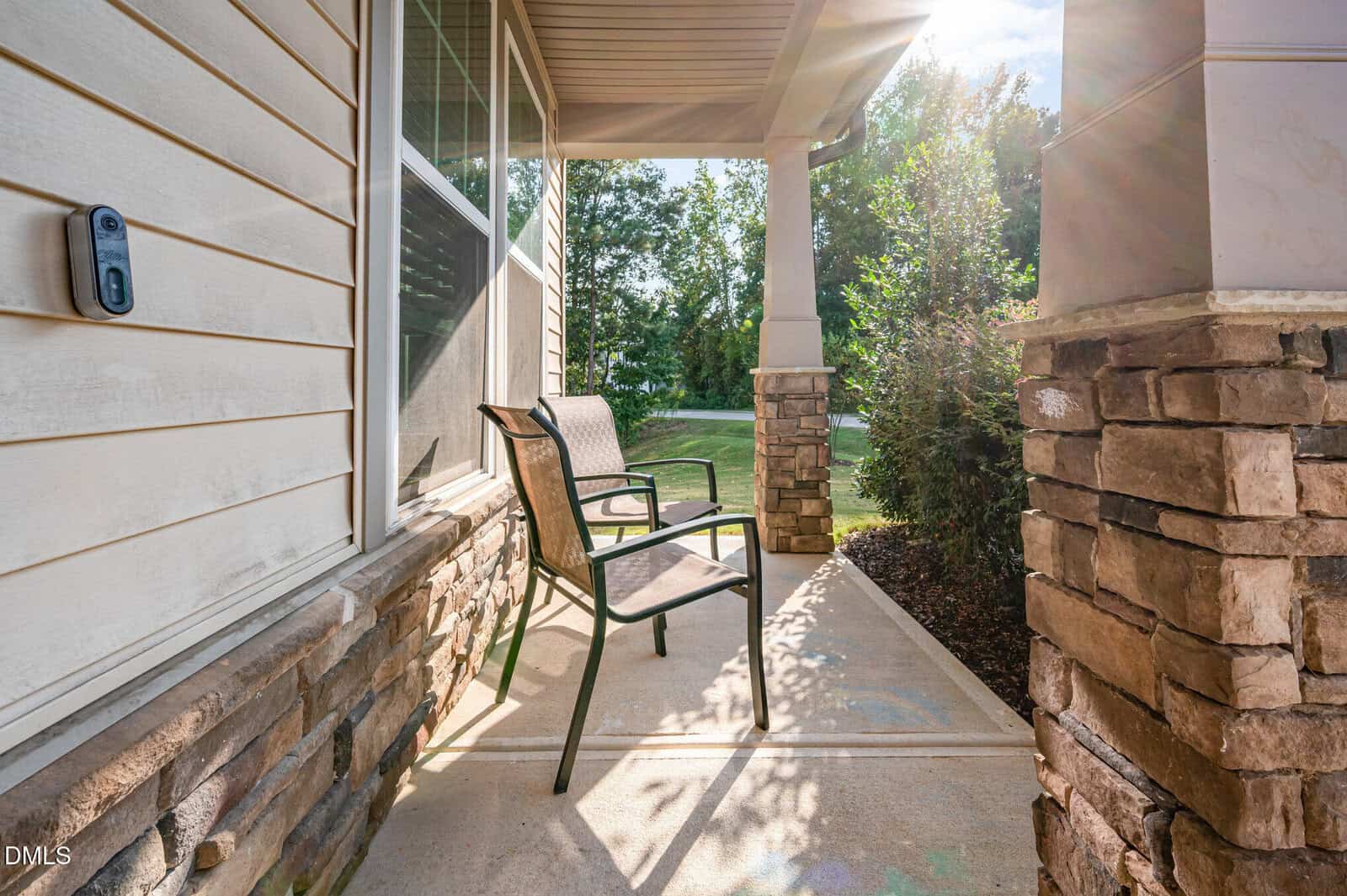 A small front porch with two metal chairs, stone pillars, beige siding, and a doorbell camera. Sunlight shines through trees, casting shadows on the concrete walkway.