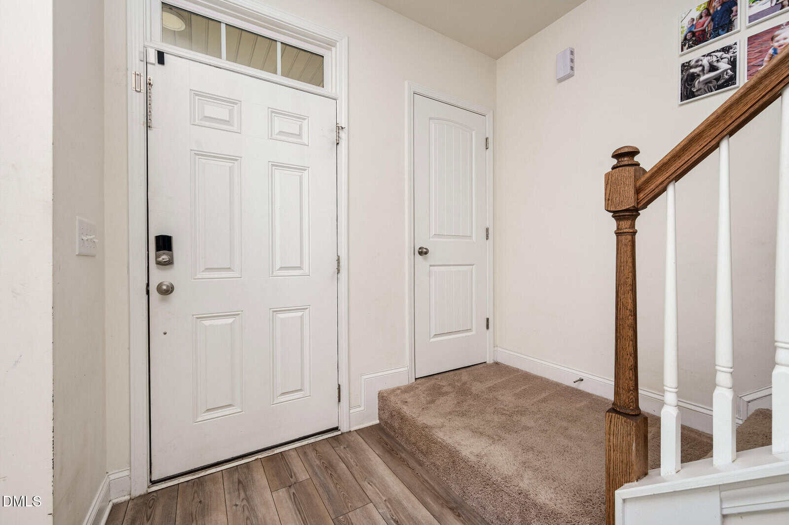A home entryway with a white front door, a second white door, carpeted stairs with a wooden handrail, light walls, wood flooring, and a few photos on the wall above the stairs.