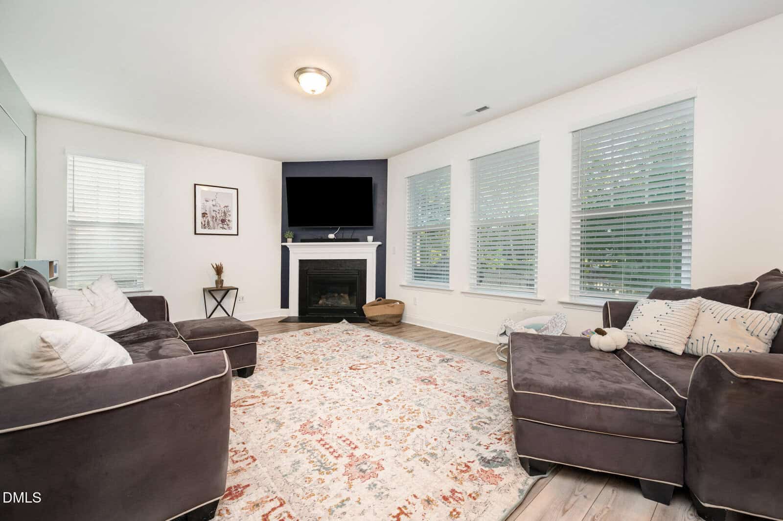 Bright living room with large windows, brown sectional sofas, patterned rug, fireplace with TV above, and light wood flooring. White walls and minimal decor create a cozy, inviting atmosphere.