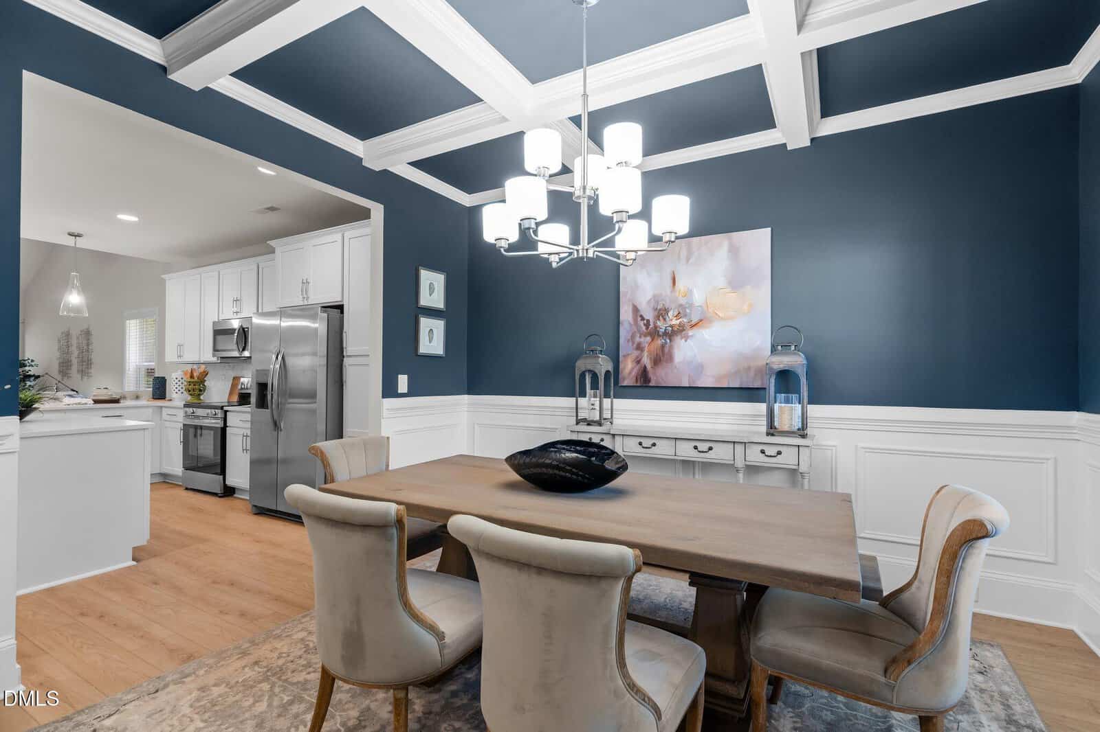 A dining room with blue walls, white coffered ceiling, chandelier, a wooden table with four upholstered chairs, decorative vase, wall art, and a view into a modern kitchen with white cabinets and stainless steel appliances.