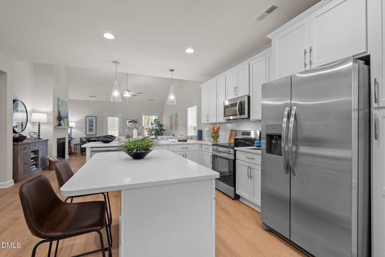 Modern kitchen with white cabinets, stainless steel appliances, an island with two brown chairs, and light wood flooring. The open layout connects to a living area with a sofa, artworks, and decor items visible in the background.