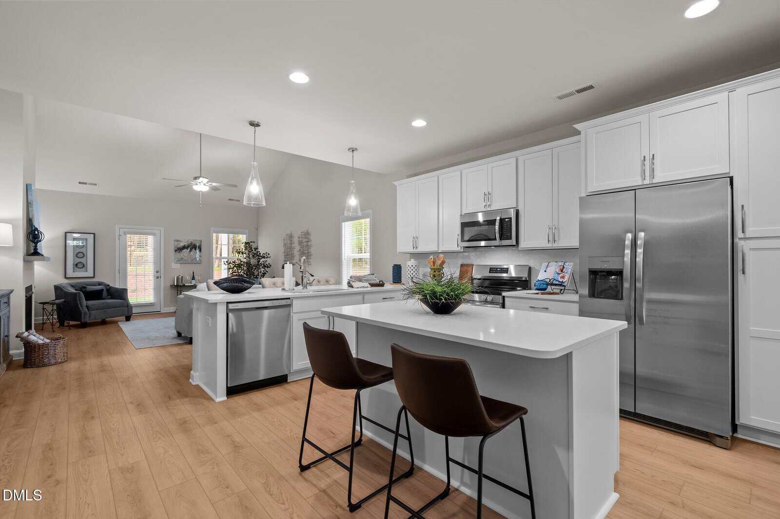 Modern kitchen with white cabinets, stainless steel appliances, and a large island with two brown chairs. Hardwood floors extend into the open living area, with visible dining and seating spaces in the background.