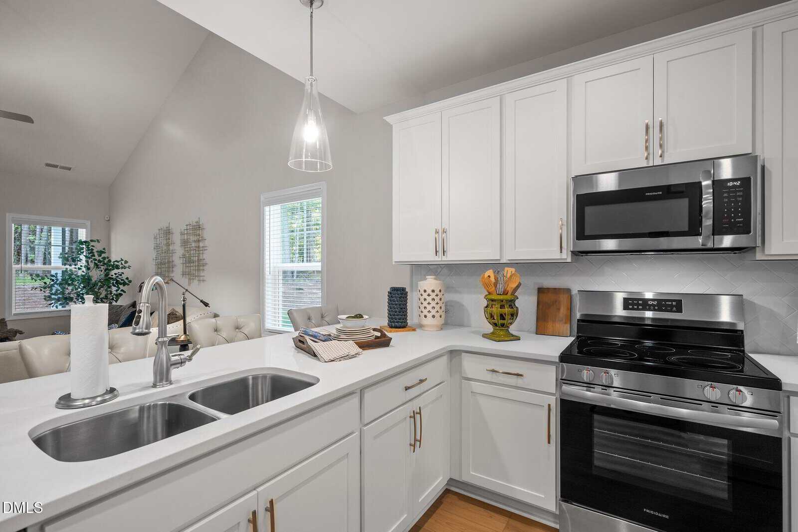 Modern kitchen with white cabinets, stainless steel appliances, double sink, and sleek fixtures. Microwave is above the stove. Countertops have decorative items, a paper towel roll, and a wooden cutting board. Dining area in background.