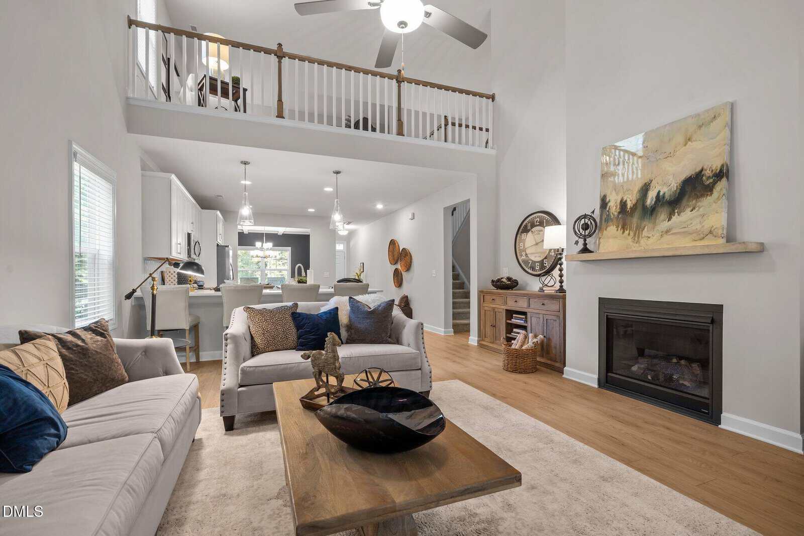 Open-concept living room with light-colored walls, two sofas, a wooden coffee table, fireplace, abstract art, and kitchen in the background. Balcony and ceiling fan visible above. Neutral decor and wood flooring throughout.