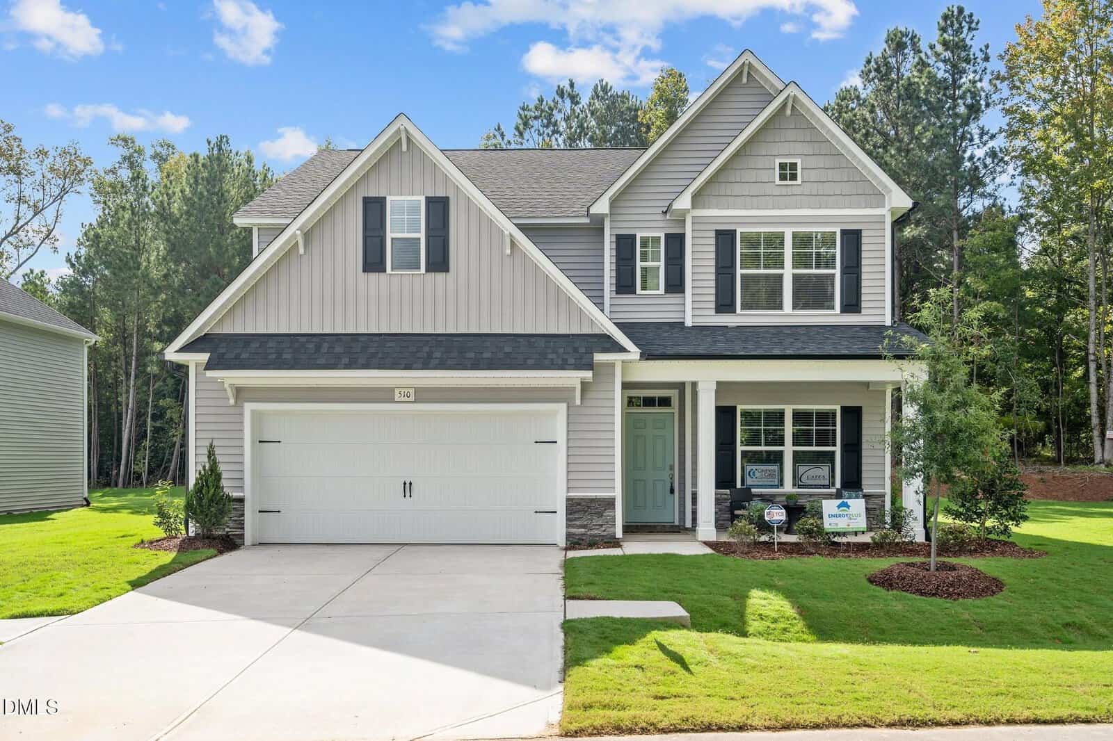 Two-story suburban house with gray siding, white trim, a double garage, green lawn, and a small tree in front. There are signs in the yard and a forested area in the background under a partly cloudy sky.