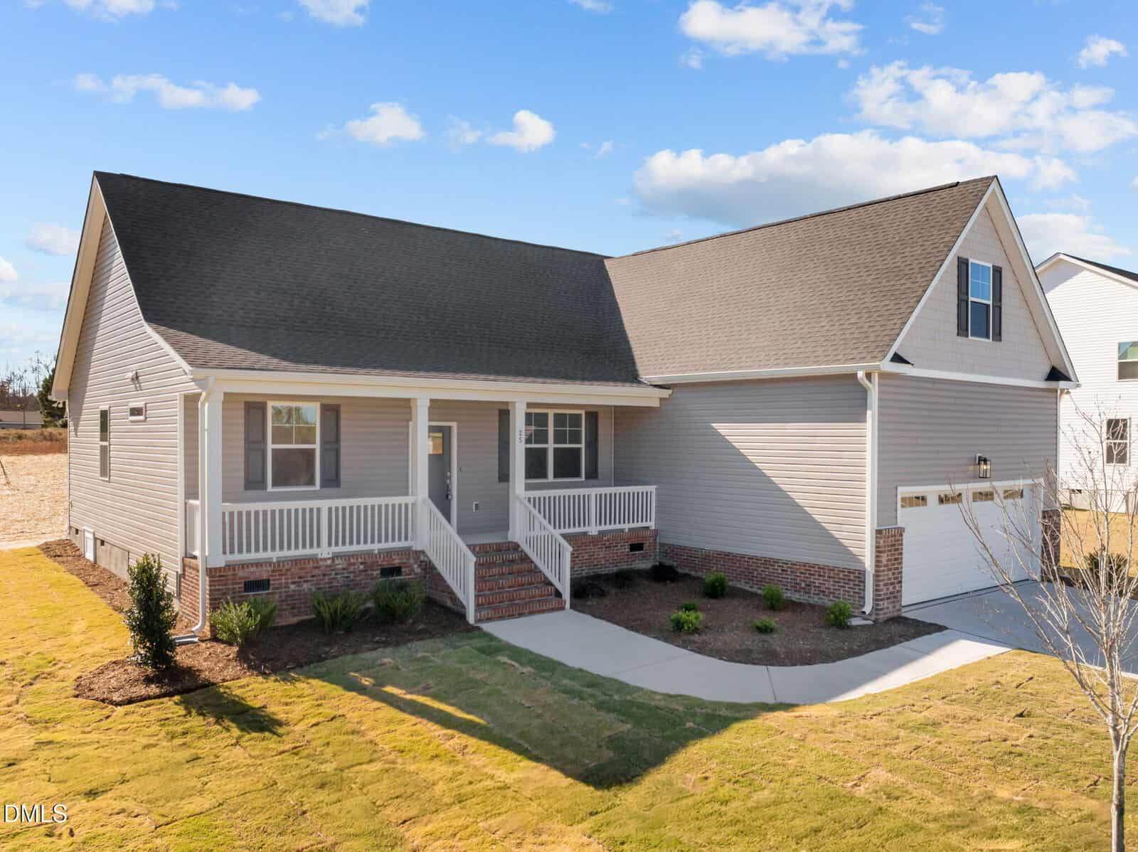 A modern single-story house with light gray siding, dark roof, attached two-car garage, covered front porch with white railings, landscaped yard, and a clear blue sky with scattered clouds.