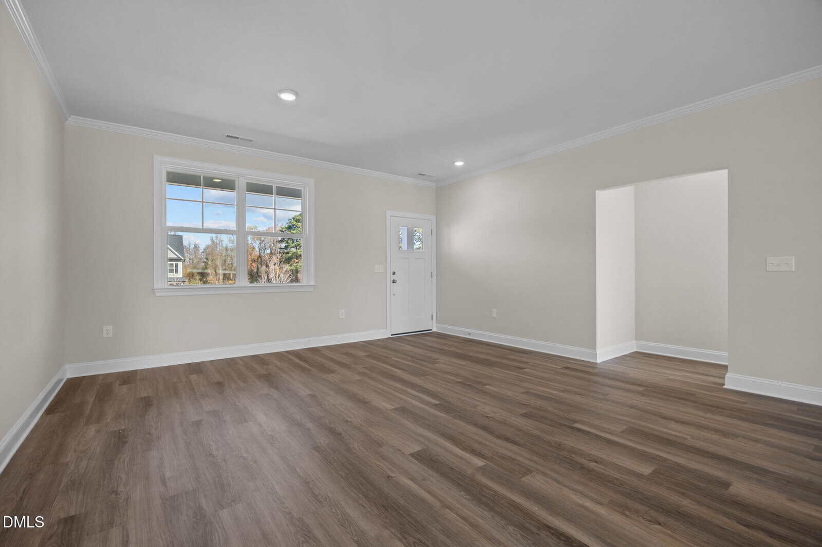 Spacious, empty living room with light beige walls, large window, white trim, wood-style flooring, recessed lighting, and a door leading outside. An open doorway leads to another room.