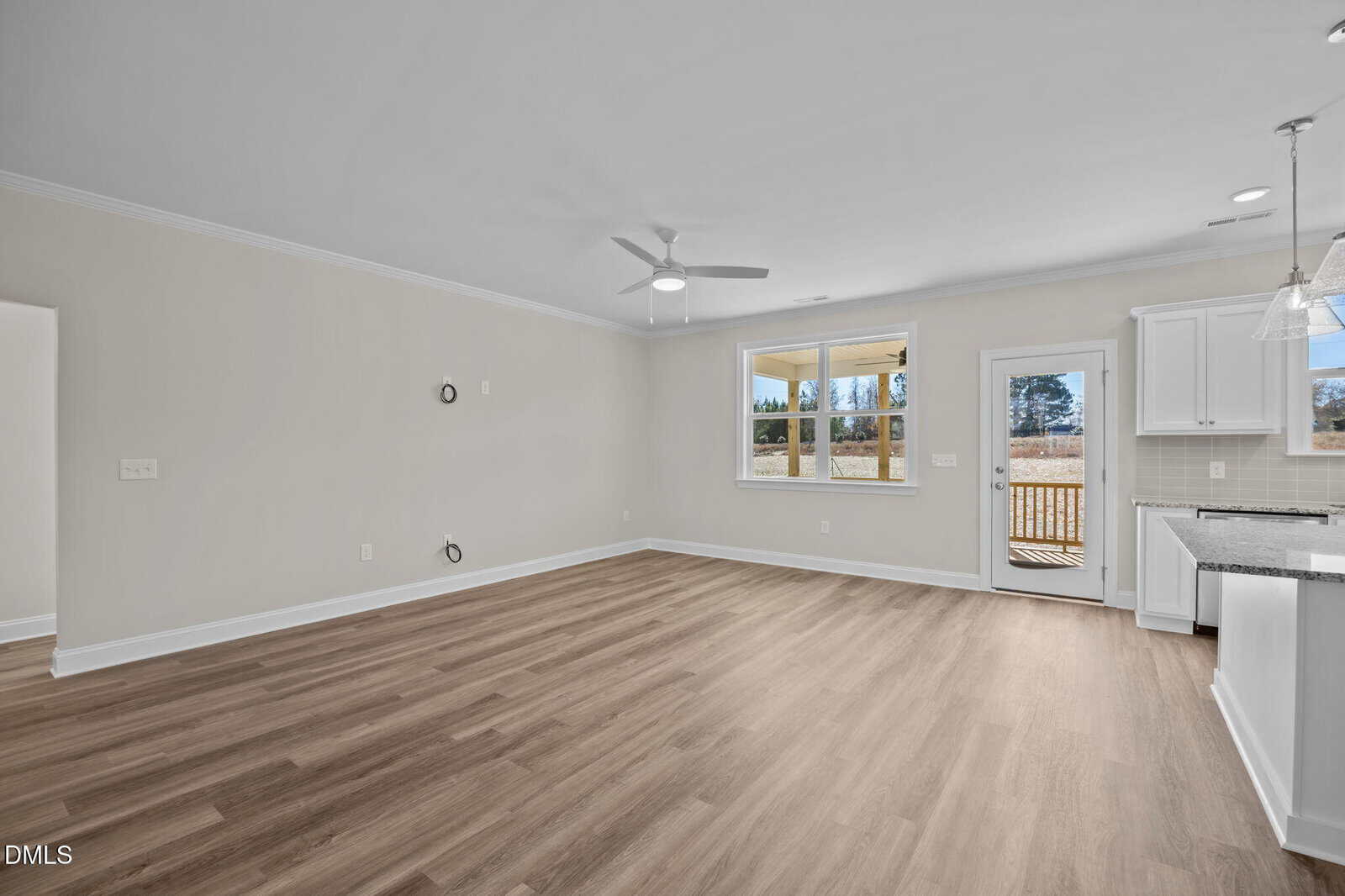 A bright, empty living room features light wood floors, beige walls, and a ceiling fan. Large windows and a glass door lead to an outdoor deck. The kitchen with white cabinets is visible to the right.