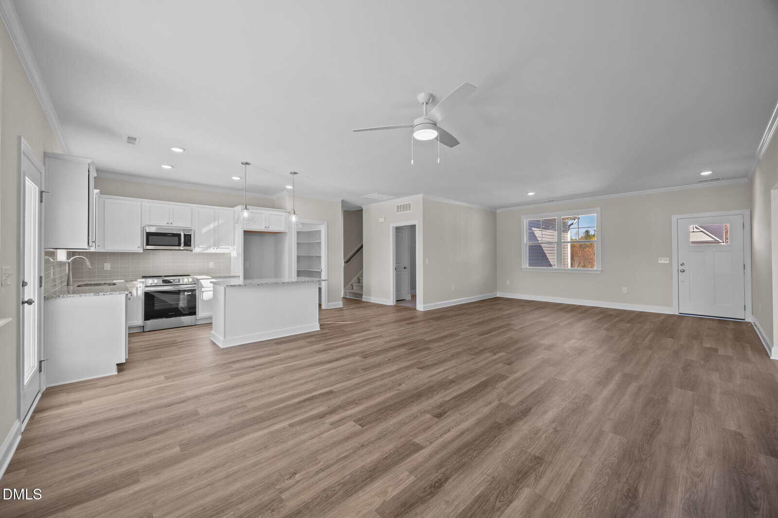 Spacious open-concept living area with wood flooring, white kitchen cabinets, modern appliances, island, ceiling fan, and windows letting in natural light. Stairs lead up, and a door is visible to the right.