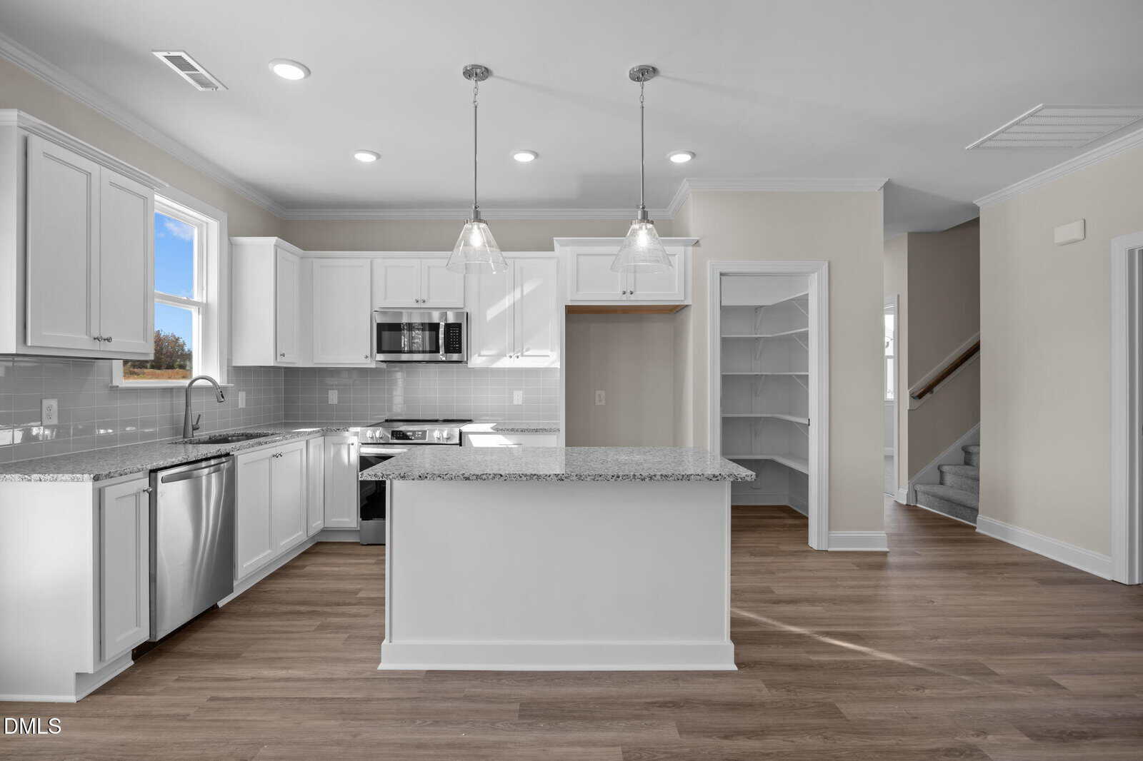 Modern kitchen with white cabinets, stainless steel appliances, granite countertops, a large center island, pendant lights, wood flooring, and an open pantry next to carpeted stairs leading to the upper floor.