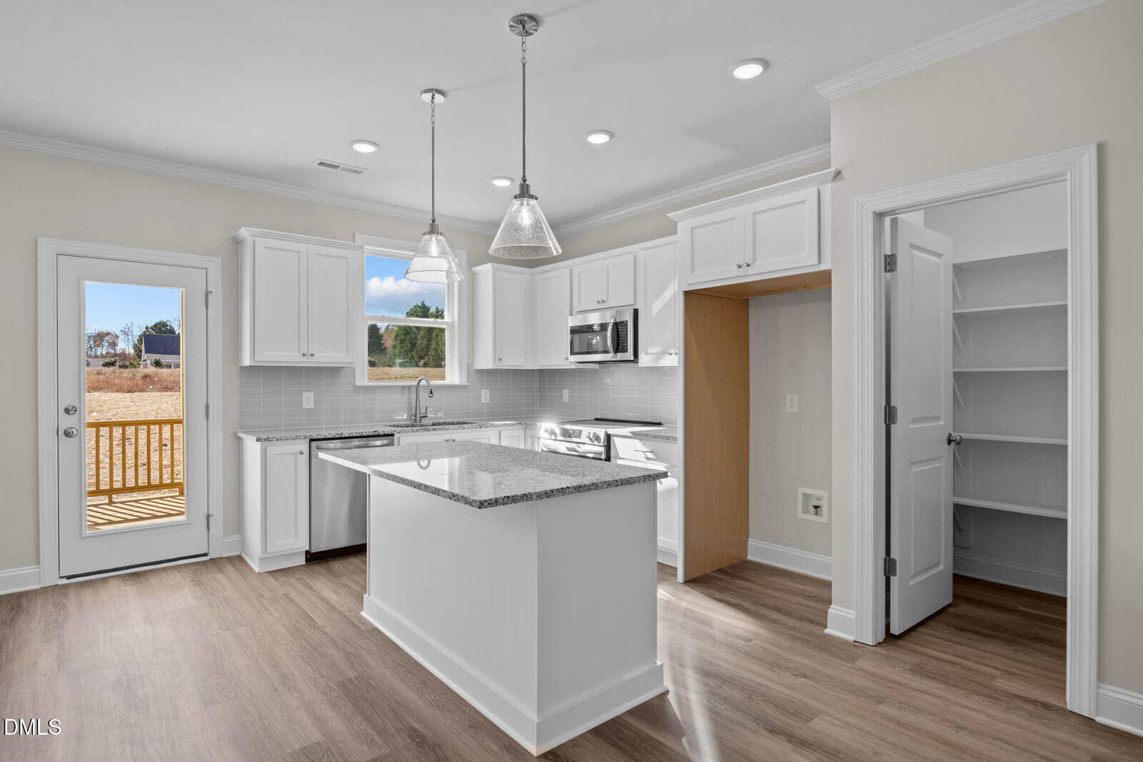Modern kitchen with white cabinets, granite countertops, an island, stainless steel appliances, pendant lighting, a walk-in pantry, and a door leading to a wooden deck. Light wood flooring and neutral walls throughout.
