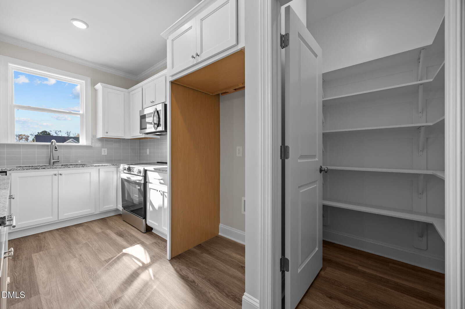 Bright modern kitchen with white cabinets, stainless steel appliances, a window, wood flooring, an empty alcove for a fridge, and an open pantry with white shelves and a door.