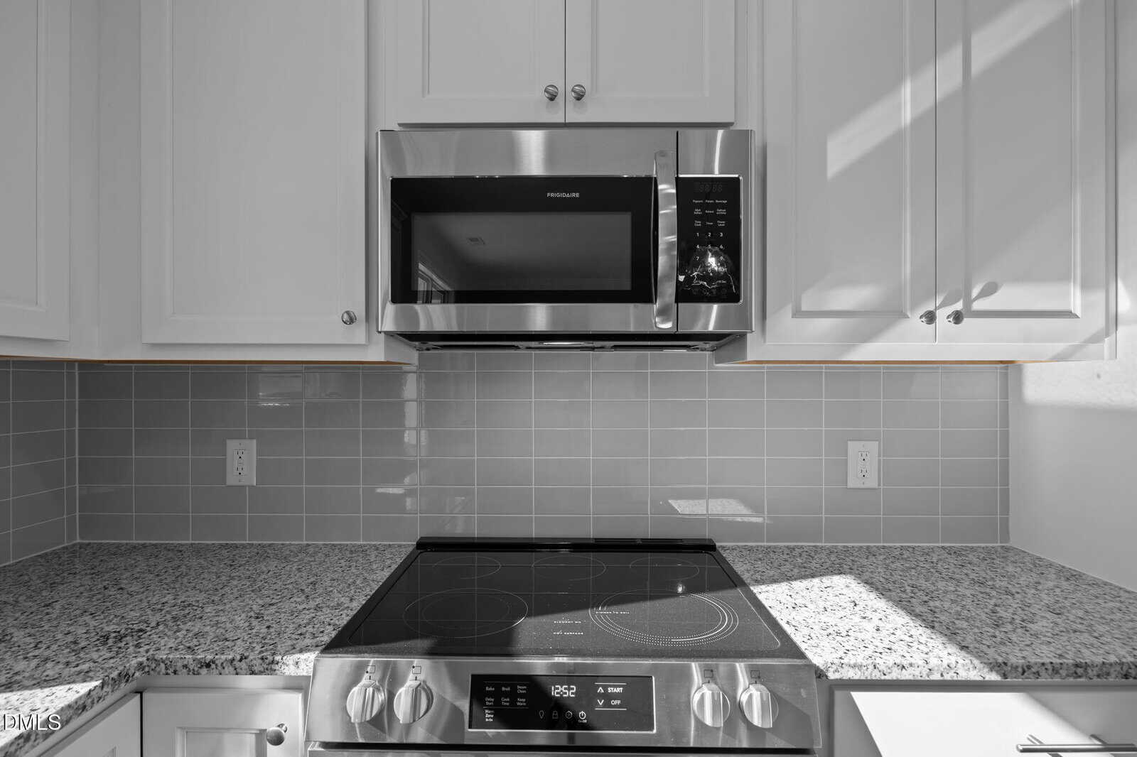 A modern kitchen features a stainless steel microwave above a matching electric stove with a glass cooktop, set against a light gray tiled backsplash and white cabinets, with sunlight streaming onto the counter.