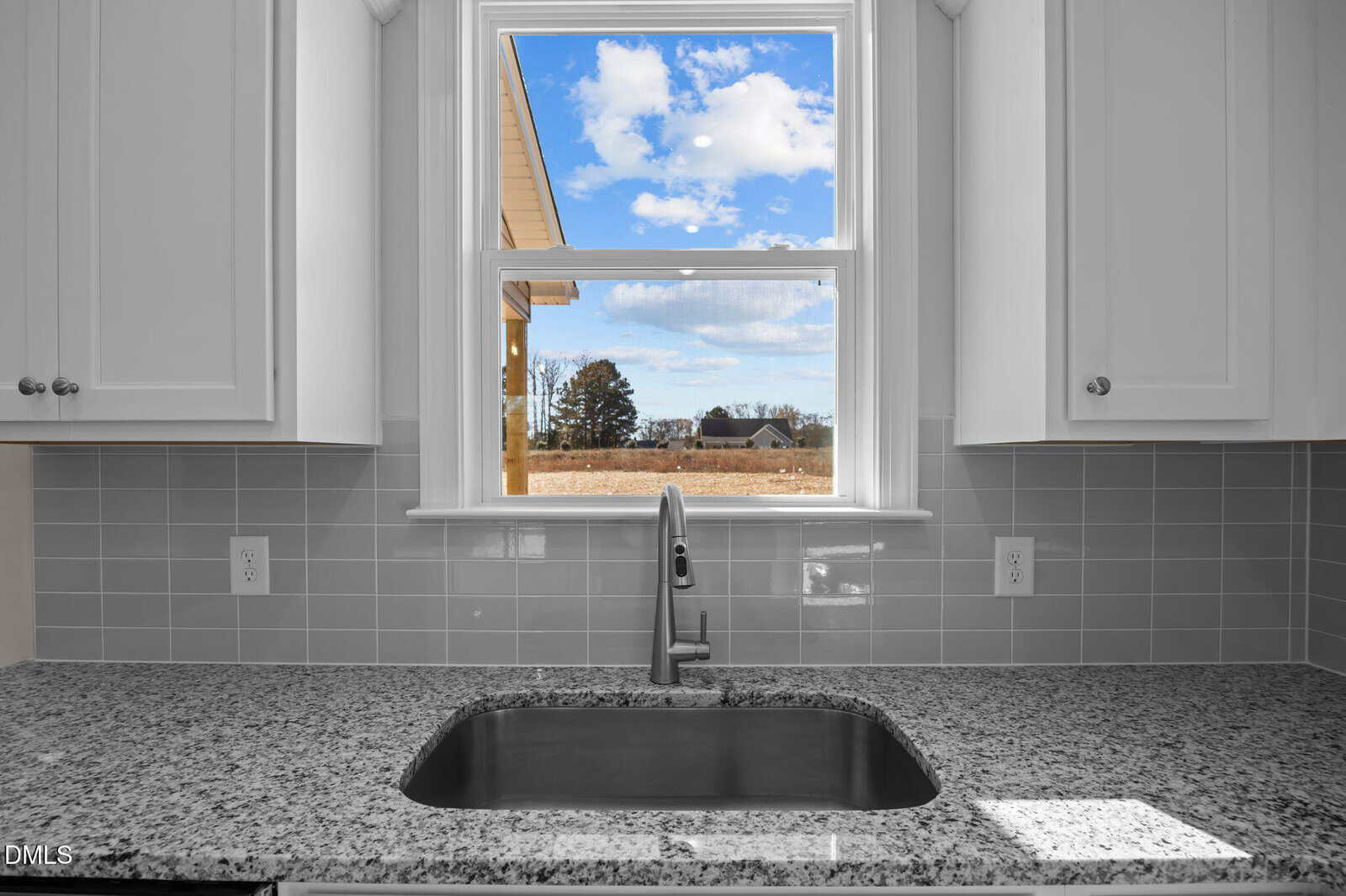A kitchen sink with a stainless steel faucet is set in a granite countertop beneath a window. Gray tile backsplash and white cabinets frame the window, which shows a clear sky and a field with houses in the distance.
