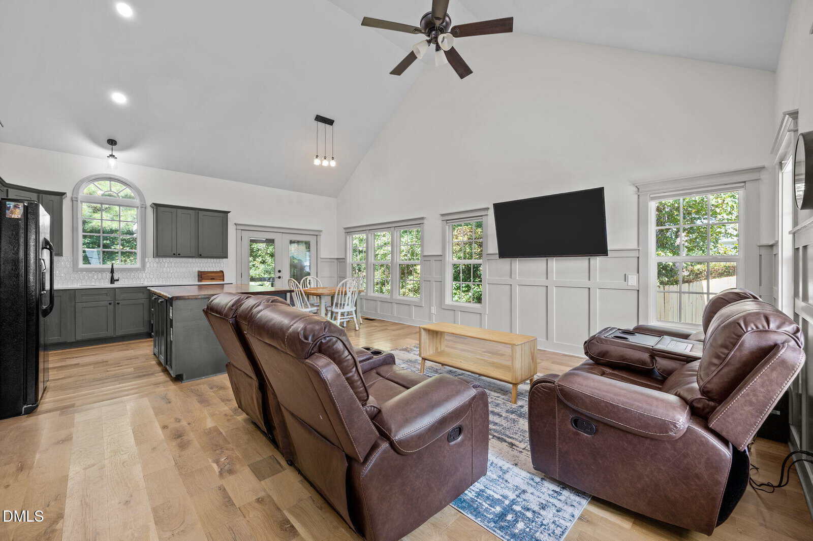 Open living area with two brown recliners, a wooden coffee table, a large wall-mounted TV, and a kitchen with gray cabinets. Multiple windows provide natural light. Light wood floors and white walls create a bright atmosphere.