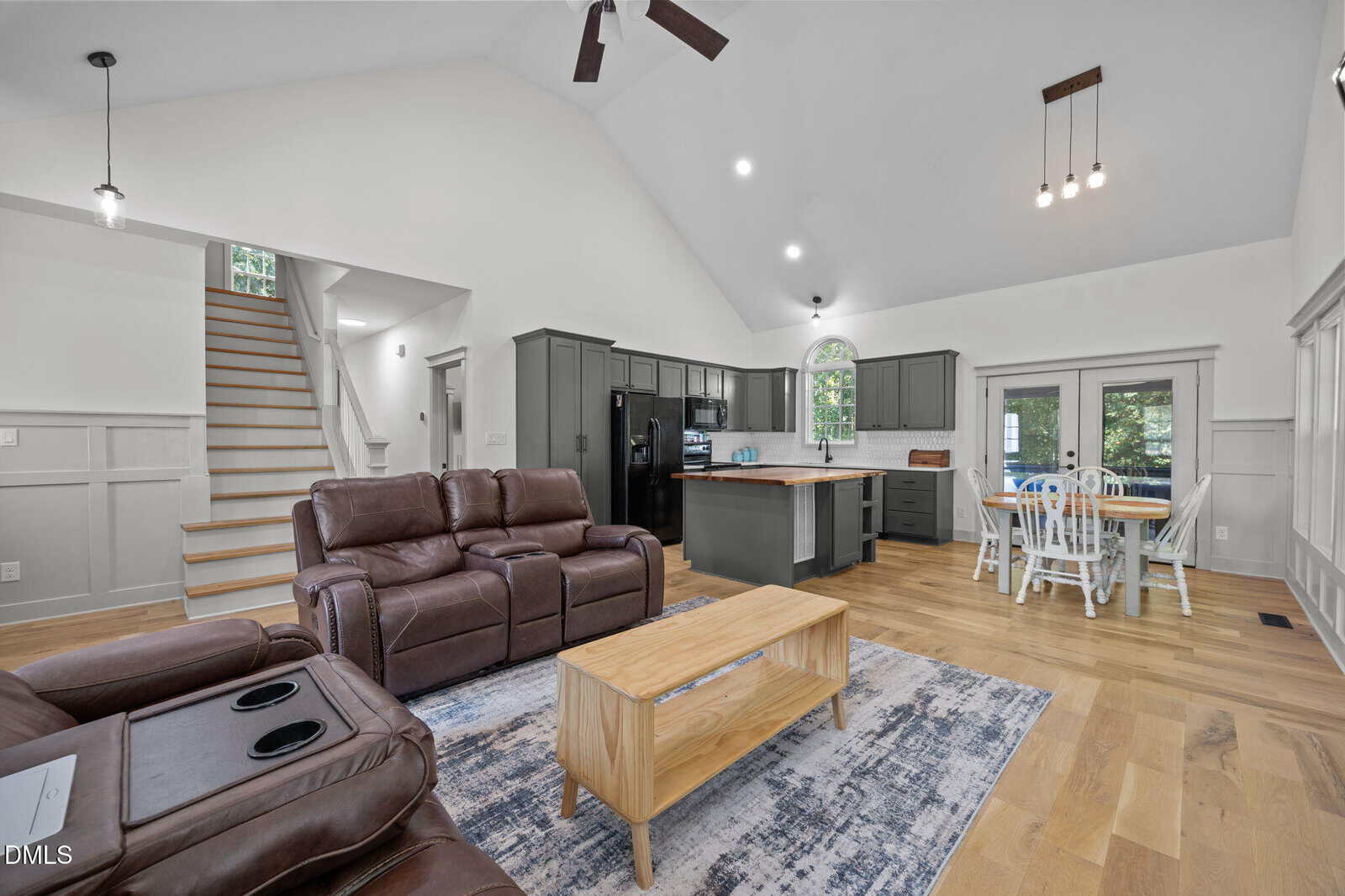 Open-concept living area with brown leather sofas, a wooden coffee table, and a rug. The kitchen has gray cabinets, an island, and a dining area with a white table and chairs. Wooden stairs lead to an upper floor.