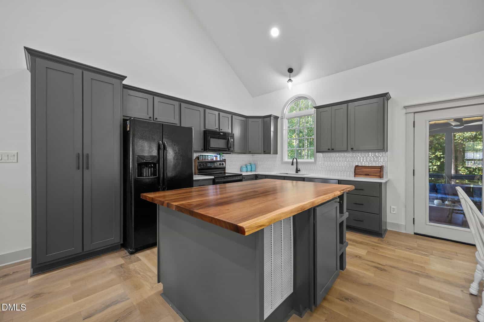 Modern kitchen with gray cabinets, black appliances, a wooden island countertop, white subway tile backsplash, and light wood flooring in 20 Richmond Place. A window above the sink lets in natural light. The dining area is visible to the right, making this Youngsville home perfect for both everyday living and entertaining.