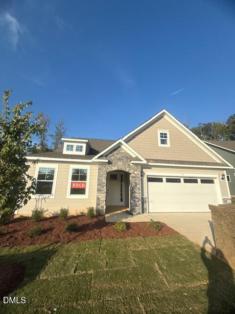 A modern, single-story house with beige siding, stone accents, a white garage door, landscaped yard, and a red "SOLD" sign in the front window, under a clear blue sky.