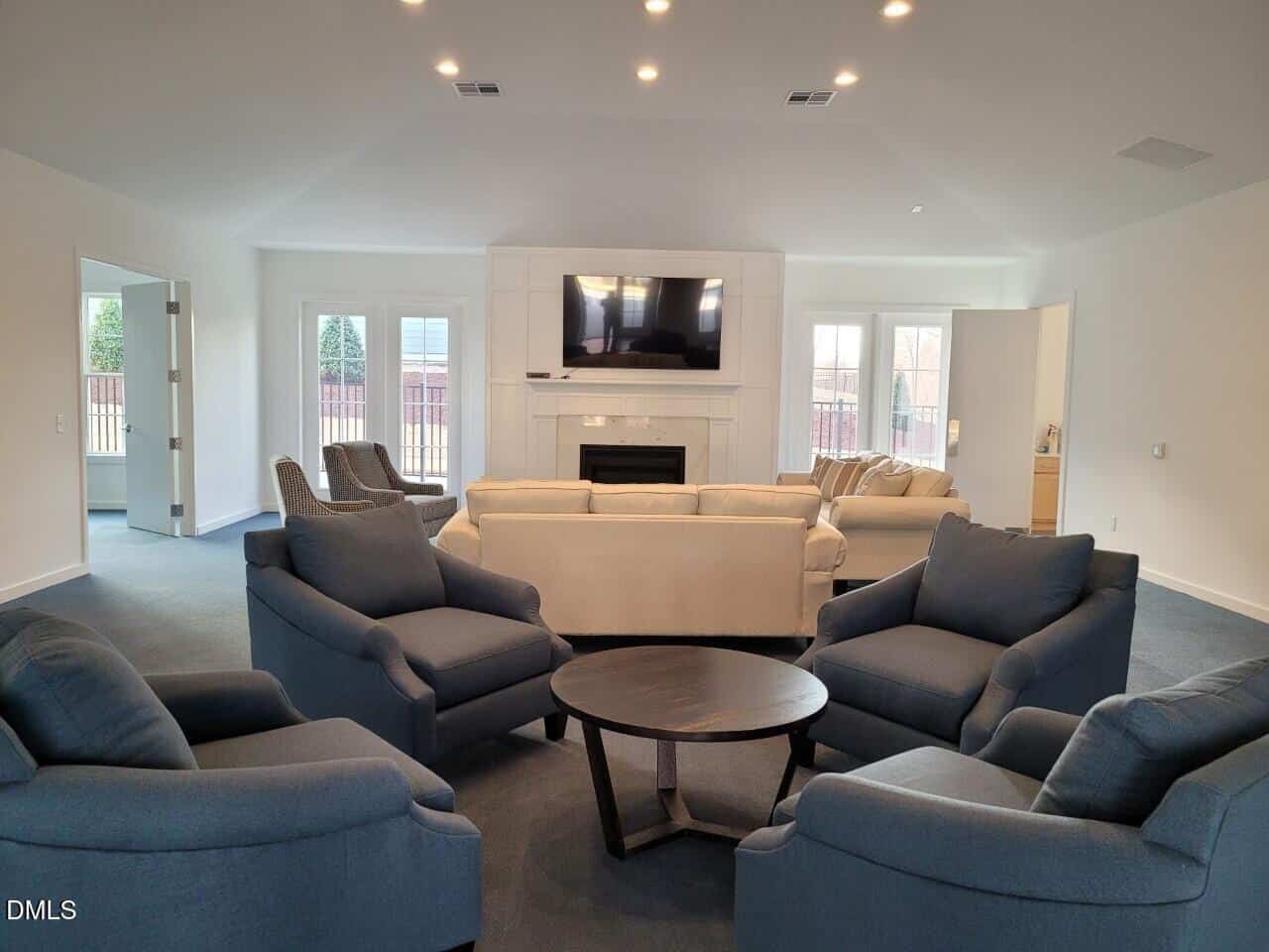 A modern living room with two beige sofas, four blue armchairs around a round black coffee table, a fireplace, and a large TV mounted above it. Large windows and glass doors let in natural light.