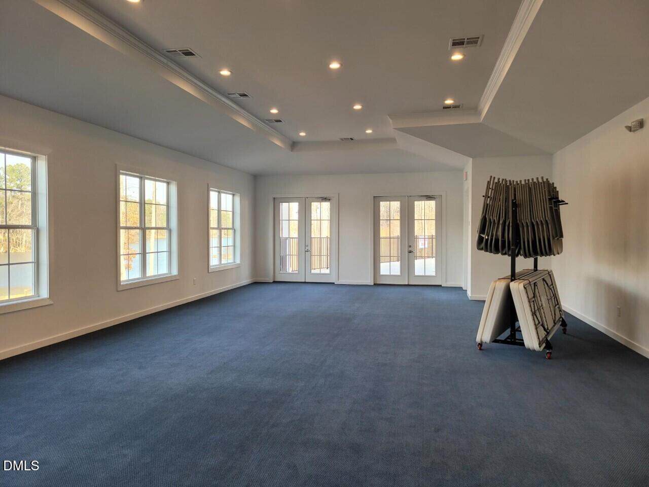 A spacious, empty room with blue carpet, white walls, large windows on one side, and double glass doors at the far end. Folded tables and stacked chairs are placed against one wall. Ceiling lights are evenly spaced overhead.