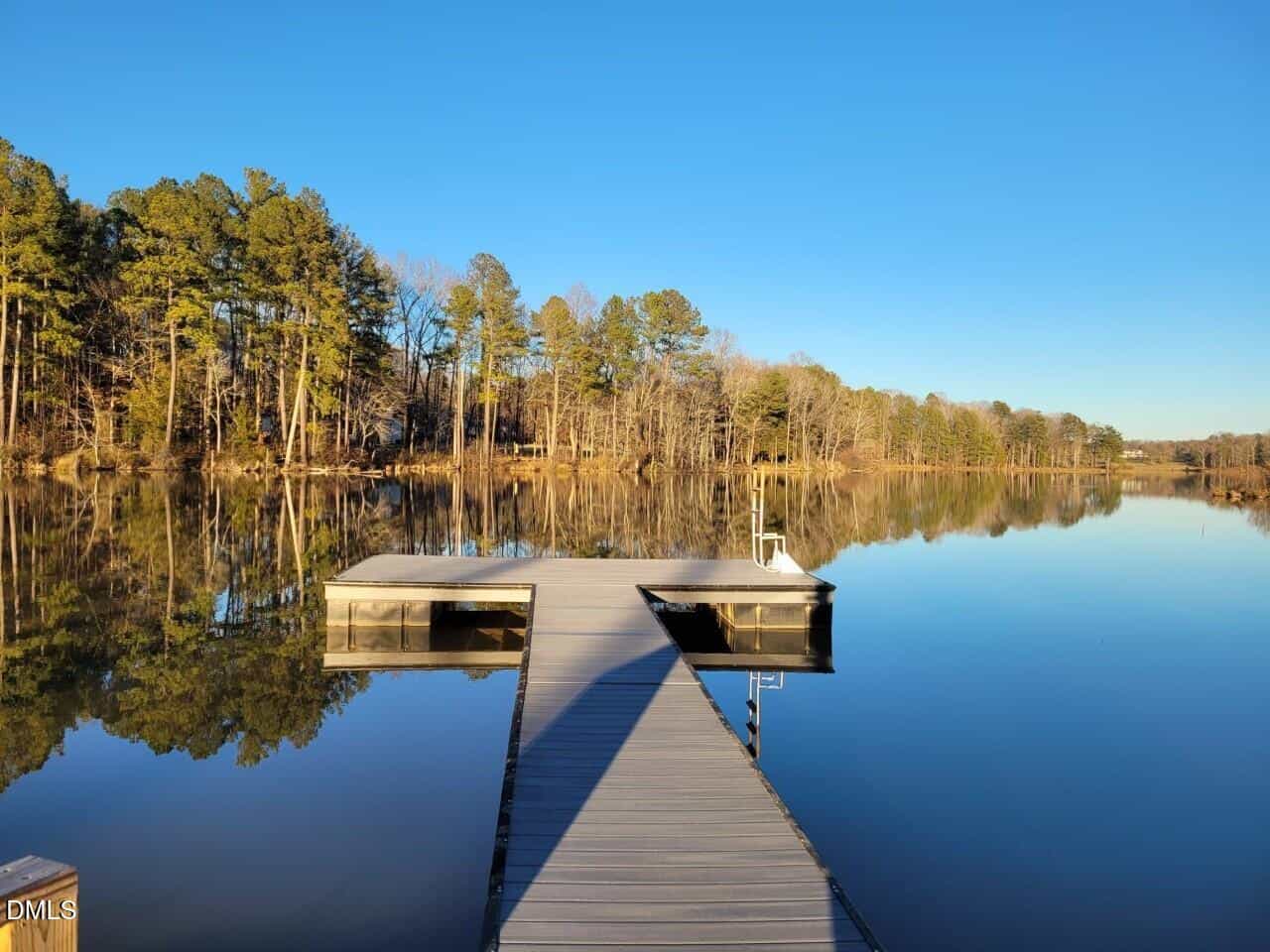 A wooden dock extends over a calm lake, reflecting the blue sky and surrounding trees. The shoreline is lined with tall trees, some with leaves and some bare, under clear daylight.