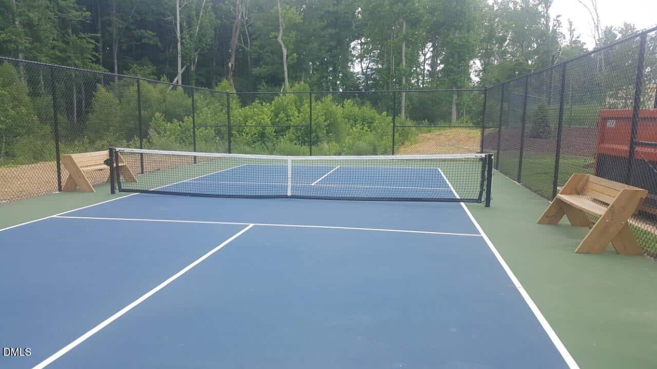 An empty outdoor tennis court with a blue surface, surrounded by a black chain-link fence, benches on each side, and trees in the background.