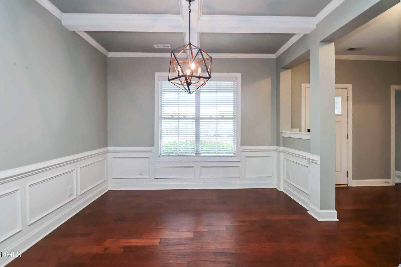 A dining room with gray walls, white wainscoting, and a modern geometric light fixture. The room has a large window with white blinds and dark wood flooring. An entryway is visible to the right.