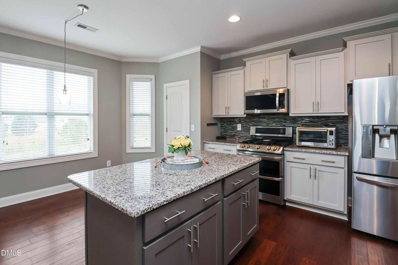 Modern kitchen with gray cabinets, stainless steel appliances, granite island countertop, wood floors, a tray with a flower vase, large windows, and neutral wall colors.