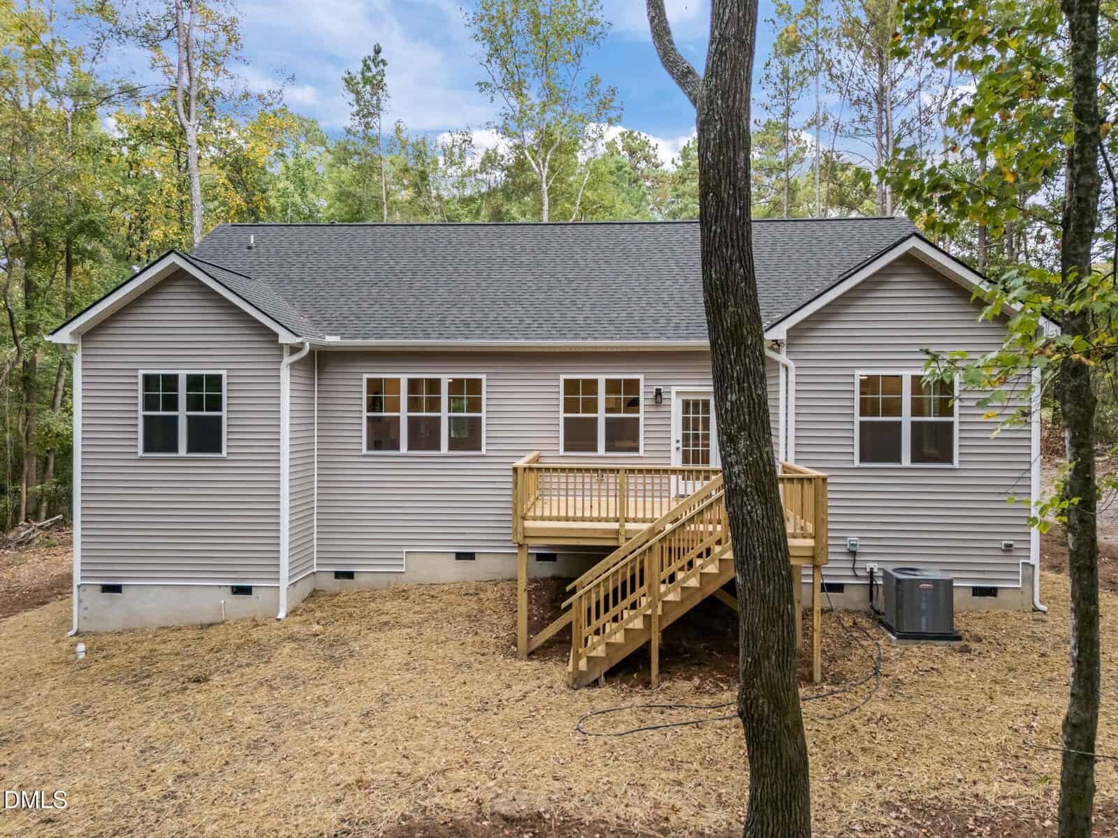 A single-story beige house with vinyl siding, multiple windows, and a small wooden deck with stairs leading to a backyard. The house is surrounded by trees and sits on a recently cleared lot.