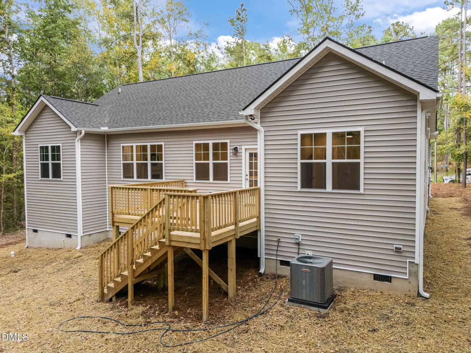 A beige single-story house with a gray shingled roof, several windows, a wooden deck with stairs, and an outdoor air conditioning unit, surrounded by trees and dirt ground.