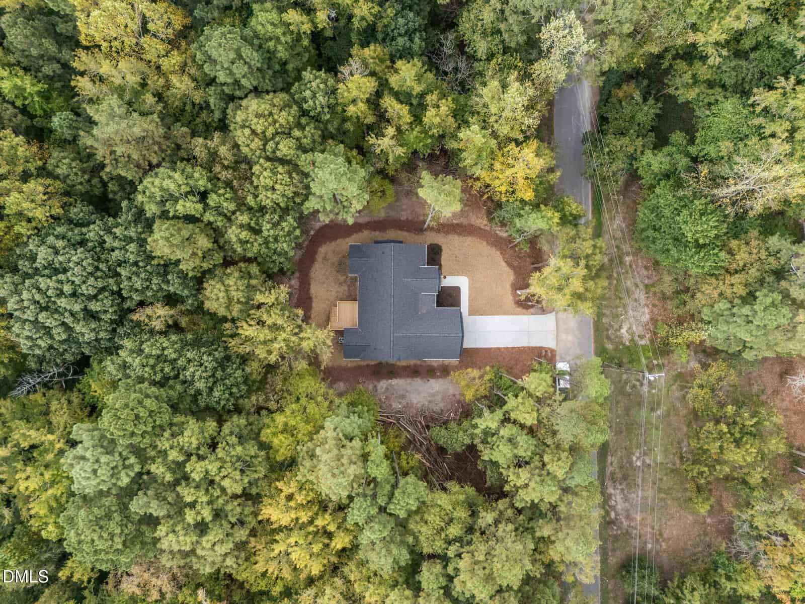 Aerial view of a single house with a gray roof, surrounded by dense green trees. The house has a driveway leading to a road, and the yard is mostly cleared with some brown patches.