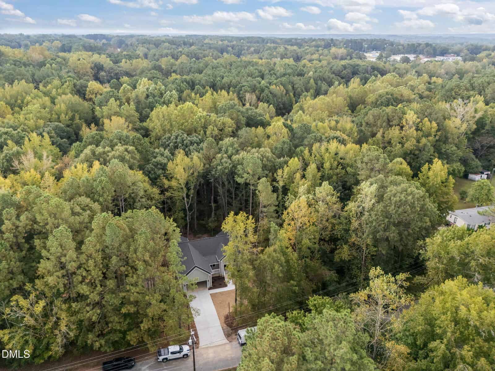 A two-story house with a driveway is surrounded by dense, green forest. The house is partially hidden by trees, and a road with a few vehicles runs along the front of the property. The sky is partly cloudy.