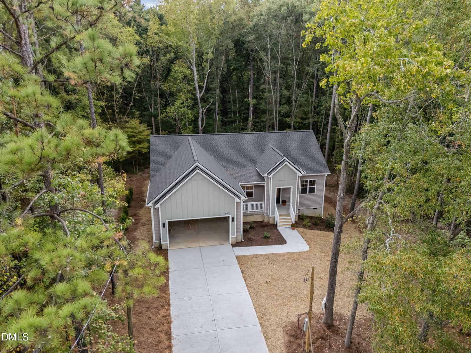 A single-story house with gray siding and a front porch sits among tall trees. The home has a gabled roof and a driveway that leads to a covered carport. The yard is partially landscaped with mulch and grass.