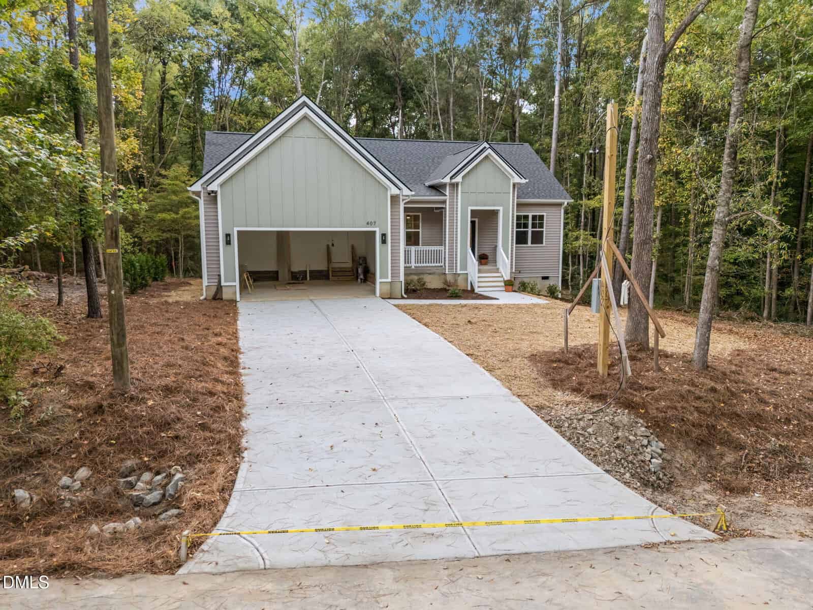 A single-story house with light green siding, a covered carport, and a front porch. The home sits at the end of a long concrete driveway surrounded by pine straw and trees on both sides.