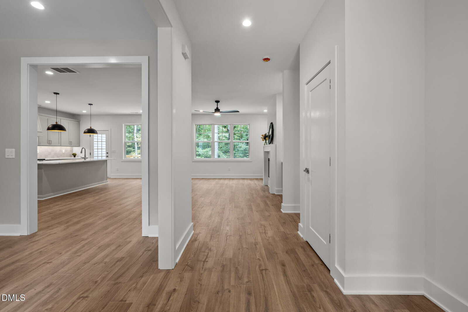 A modern home interior with light gray walls, white trim, and wood flooring. The hallway leads to an open living area with large windows, a ceiling fan, and a view of the kitchen on the left.