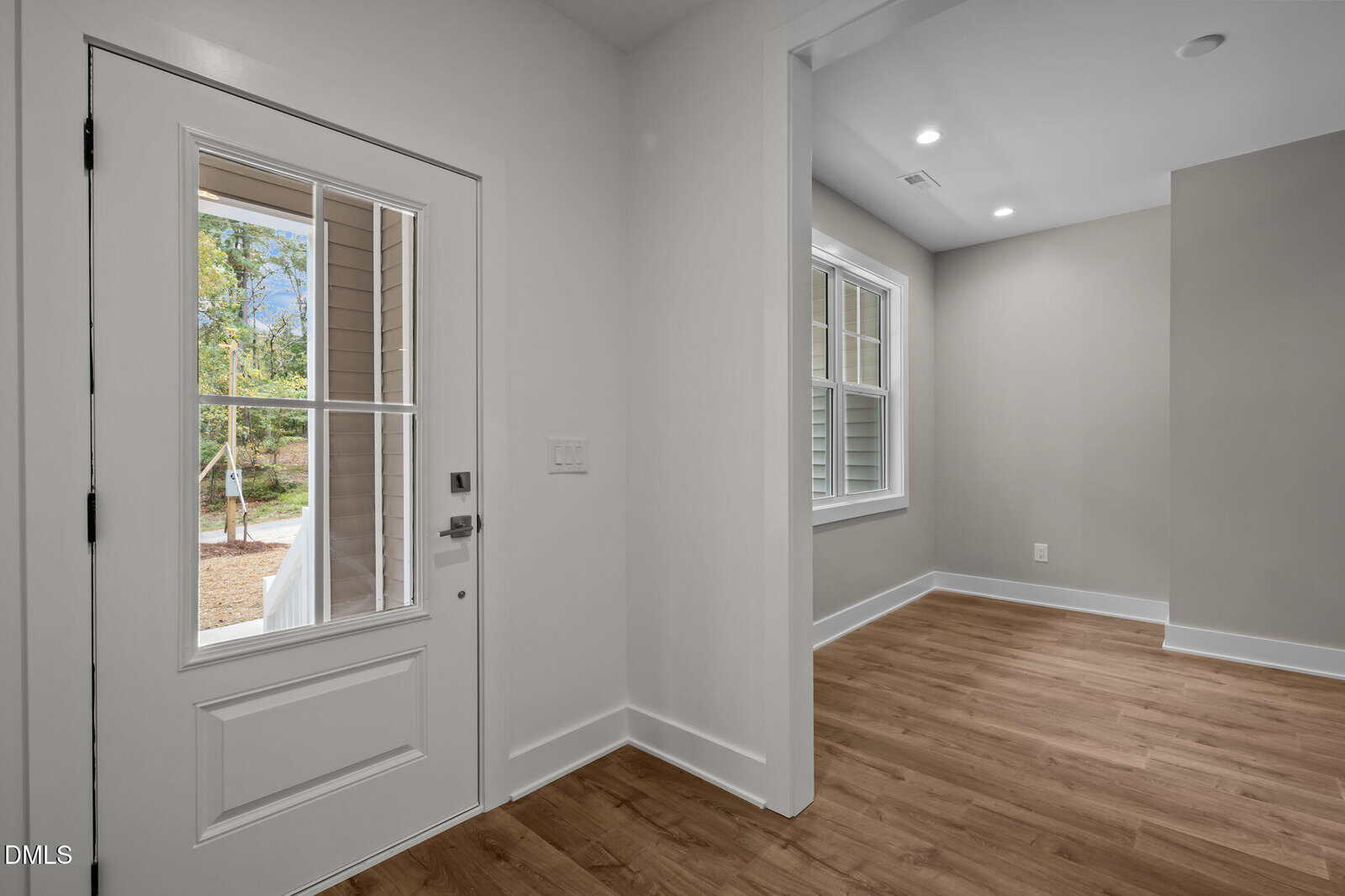 A view of a home's entryway with a white front door that has glass panels, looking into an adjacent room with large windows, light gray walls, and wood flooring. The space is well-lit with natural light.