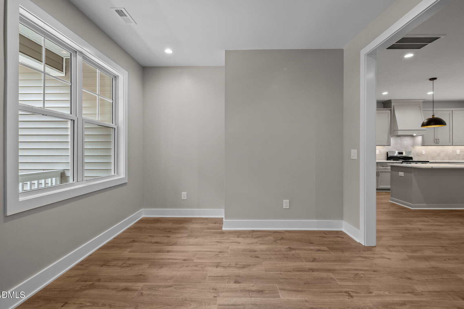 Unfurnished room with light brown wood flooring, light gray walls, a double window on the left, recessed ceiling lights, and an open doorway leading to a kitchen with white cabinets and stainless steel appliances.