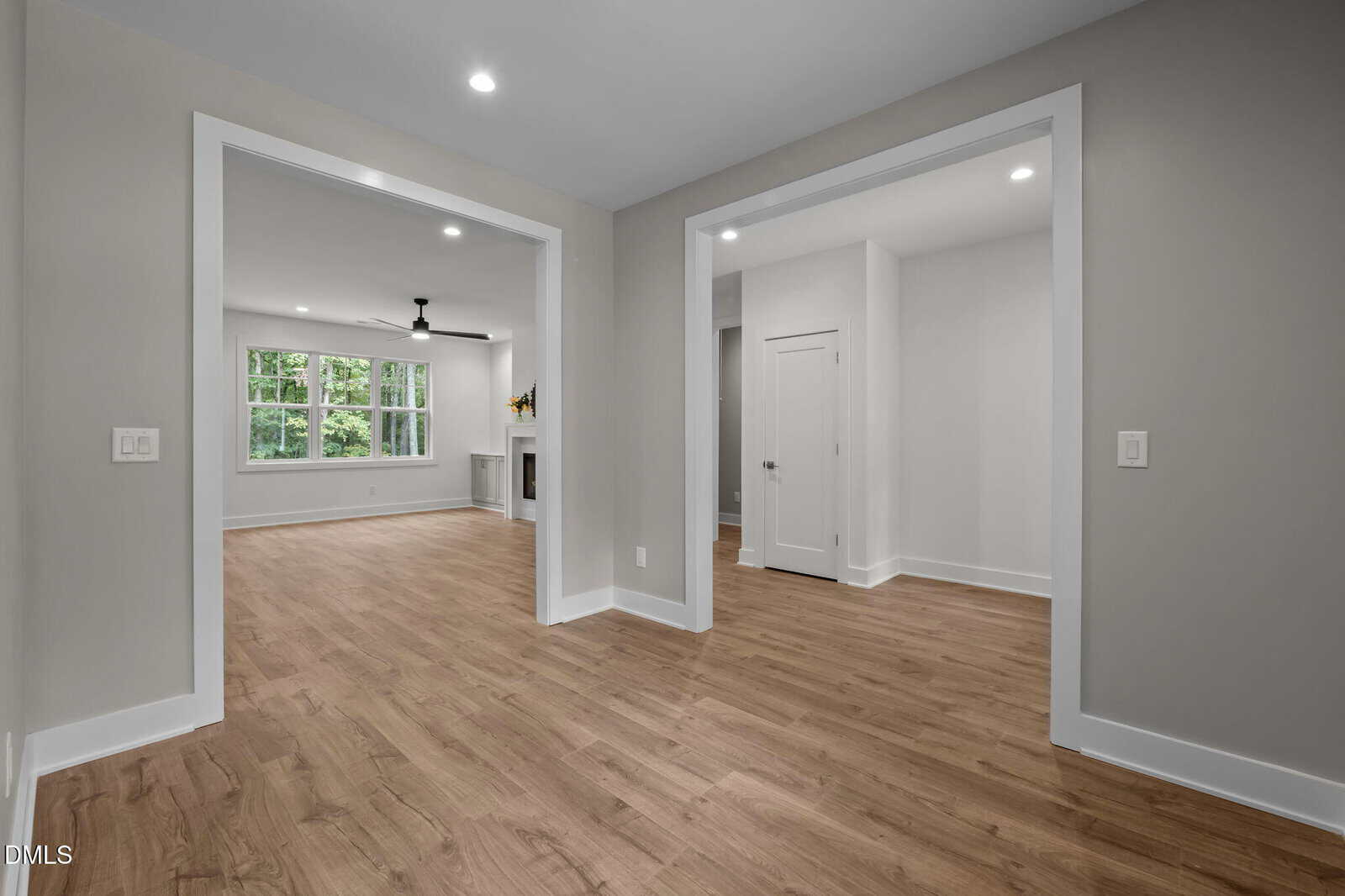 A modern interior space with light wood flooring, light gray walls, white trim, recessed ceiling lights, and large windows allowing natural light into the living area. A ceiling fan and fireplace are visible in the background.