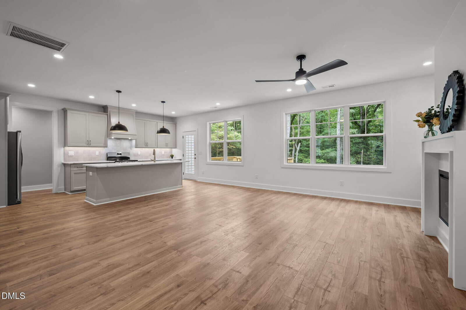 Spacious open-concept living area with wood-style flooring, modern kitchen with white cabinets and pendant lights, large windows, ceiling fan, and a fireplace; backyard view through the windows.