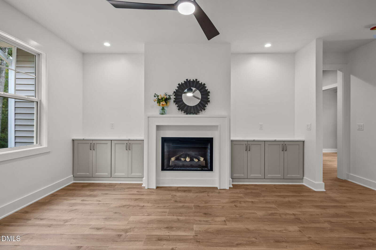 Modern living room with wood flooring, a white fireplace, gray built-in cabinets on both sides, a round mirror, vase with flowers, ceiling fan, and a window to the left.