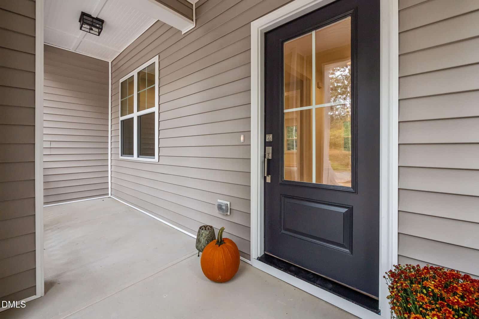 A tan house with white trim features a black front door with glass panels. A pumpkin and a decorative gourd sit on the porch near the door. A potted plant is at the edge of the image.