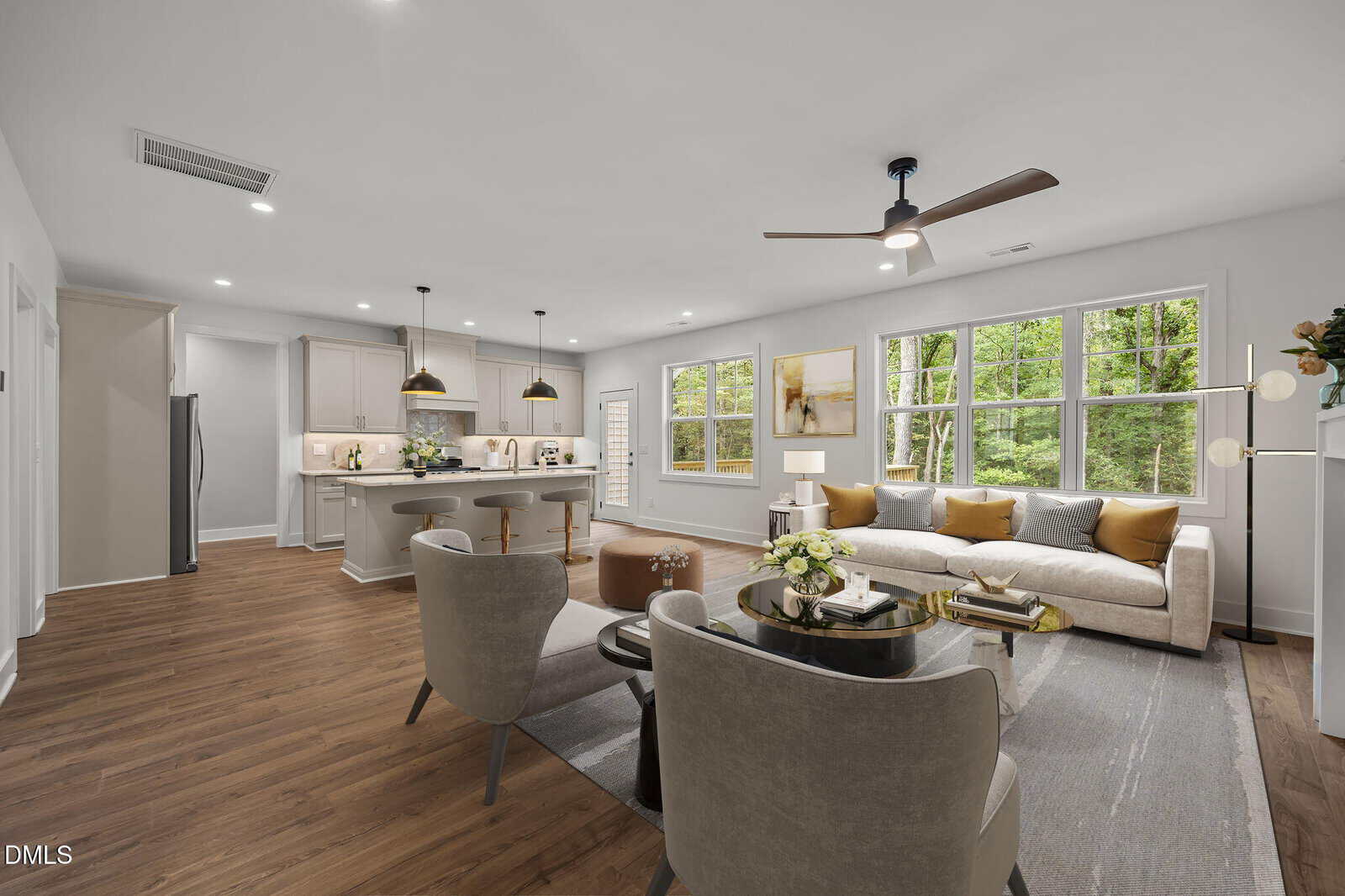 Modern open living room and kitchen, featuring wood floors, neutral furniture, large windows with forest view, and ceiling fans. Dining area with stools near kitchen island; stylish, bright, and inviting space.