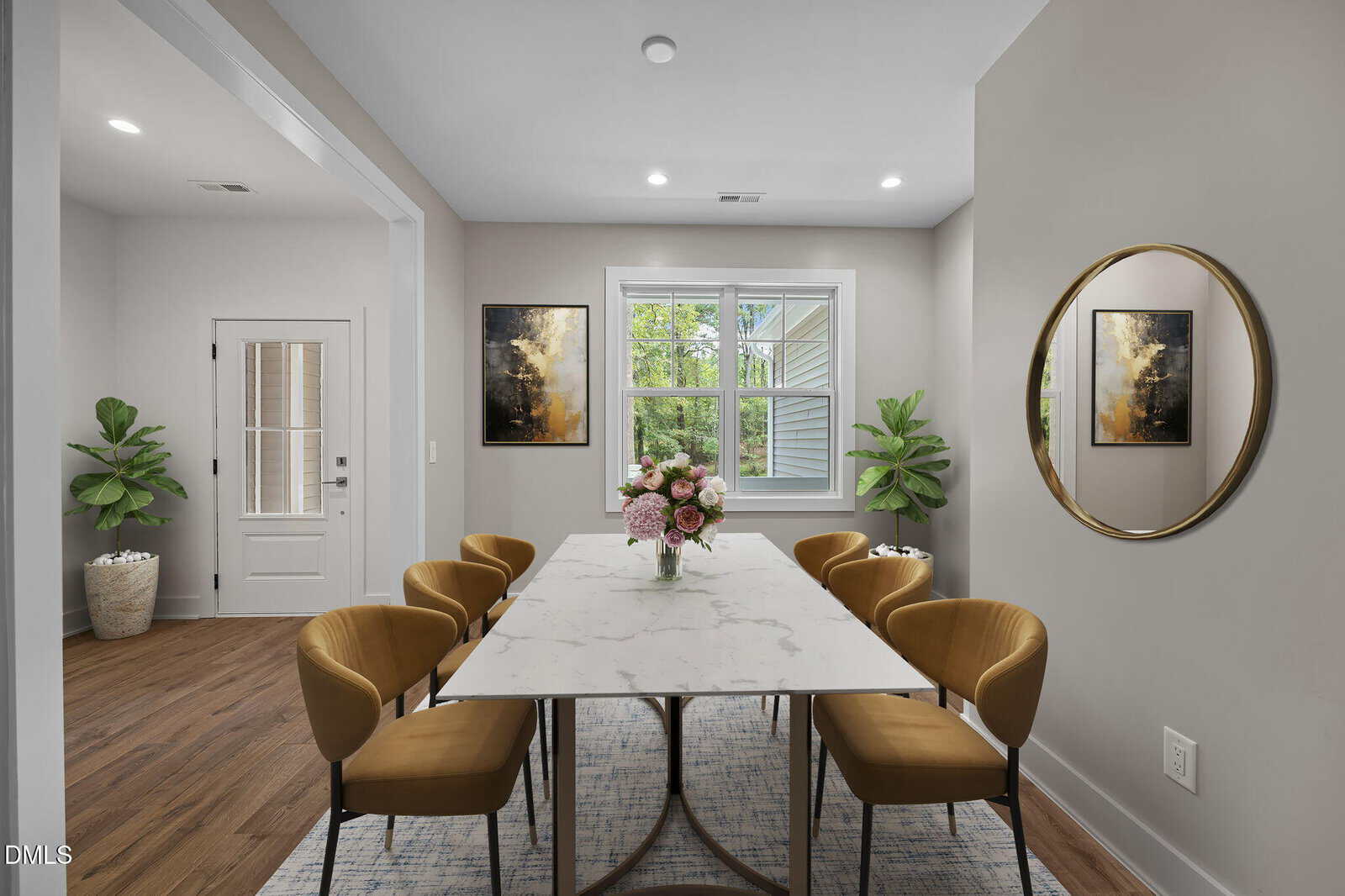 A modern dining room with a marble table, six mustard yellow chairs, a round mirror on the wall, two framed artworks, potted plants, and wood flooring. A door and window allow natural light into the space.