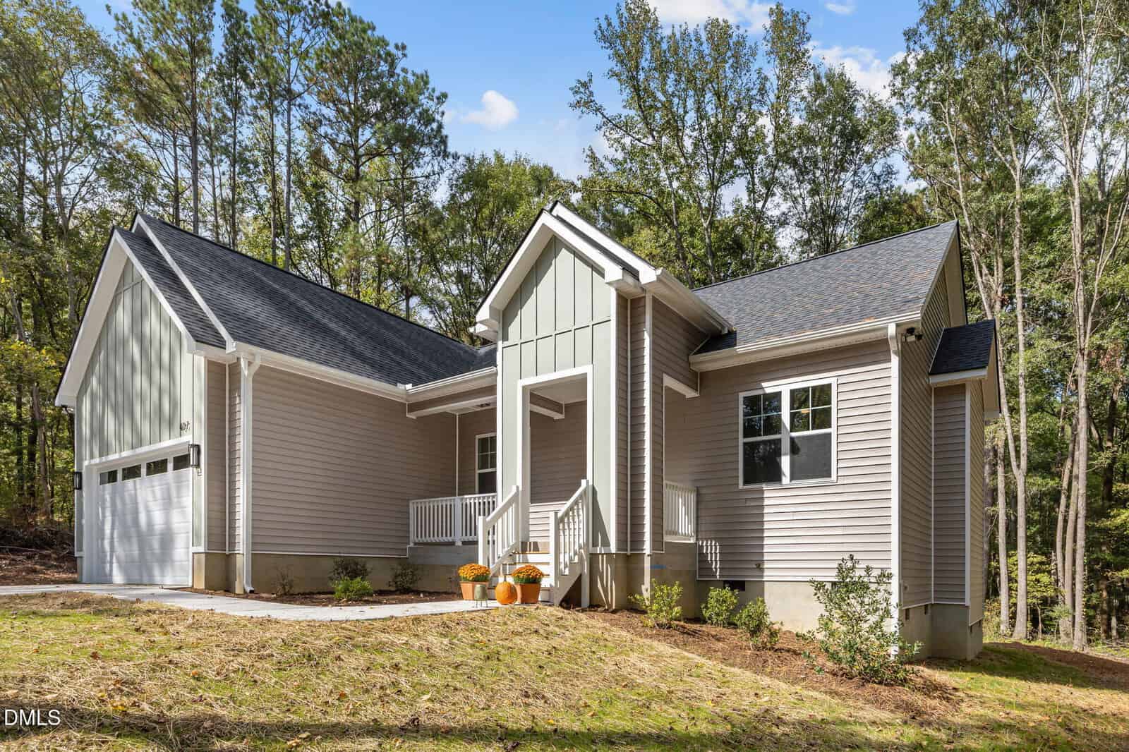 Single-story modern house with light gray siding, white trim, a dark shingled roof, and an attached two-car garage. The entrance has steps, a small porch, and two pumpkins on display. The home is surrounded by trees.