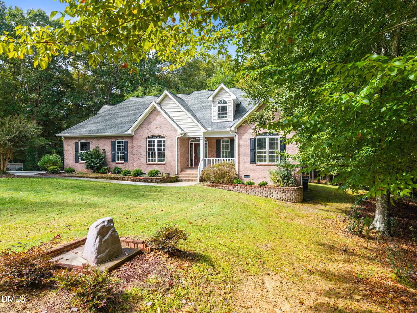 Single-story brick house with gray roof, black shutters, and white trim; front entry has steps and railings. The yard is grassy with landscaping, trees, and a stone marker near the driveway.